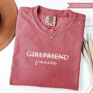 May include: Crimson-colored t-shirt with the words "GIRLFRIEND" and "fianc&eacute;e" printed in white. A gold pendant necklace rests on the shirt. A brown hat and dried decorative leaves are also in the image.