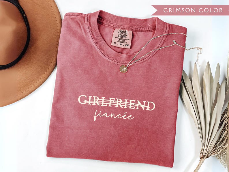 May include: Crimson-colored t-shirt with the words "GIRLFRIEND" and "fianc&eacute;e" printed in white. A gold pendant necklace rests on the shirt. A brown hat and dried decorative leaves are also in the image.