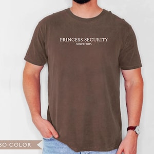 May include: Espresso-colored t-shirt with the text "PRINCESS SECURITY SINCE 2013" printed in white across the chest. The shirt is a solid color and has short sleeves. The model is wearing a watch with a brown leather band.