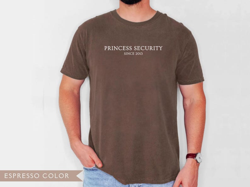 May include: Espresso-colored t-shirt with the text "PRINCESS SECURITY SINCE 2013" printed in white across the chest. The shirt is a solid color and has short sleeves. The model is wearing a watch with a brown leather band.