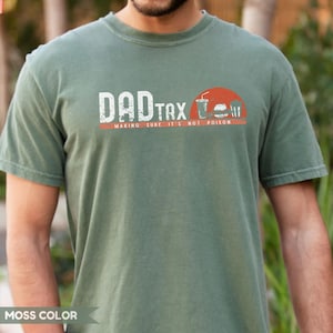 May include: Moss green t-shirt with the text "DAD TAX" in white, above a red line with the words "MAKING SURE IT'S NOT POISON". A graphic of a drink, burger, and fries is on the right.