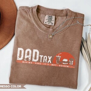 May include: Espresso-colored t-shirt with the words "DAD TAX" in white block letters and a red stripe underneath. A graphic of a drink, burger, and fries is on the right. The phrase "MAKING SURE IT'S NOT POISON" is below the text.