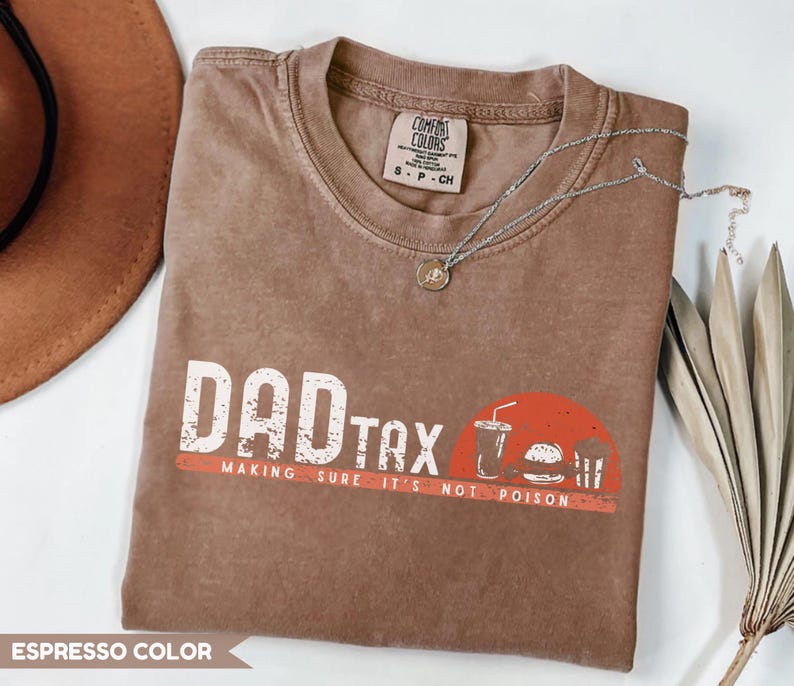 May include: Espresso-colored t-shirt with the words "DAD TAX" in white block letters and a red stripe underneath. A graphic of a drink, burger, and fries is on the right. The phrase "MAKING SURE IT'S NOT POISON" is below the text.