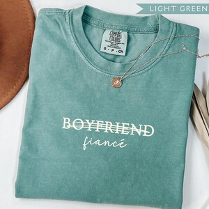 May include: A light green t-shirt with the words "BOYFRIEND" and "fianc&eacute;" printed in white. A silver necklace with a pendant rests on the shirt. A brown hat and dried plant are in the background.