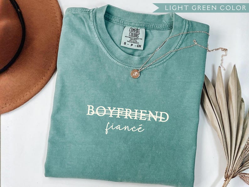 May include: A light green t-shirt with the words "BOYFRIEND" and "fianc&eacute;" printed in white. A silver necklace with a pendant rests on the shirt. A brown hat and dried plant are in the background.