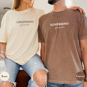 May include: Two matching t-shirts, one ivory and one espresso, with the words "GIRLFRIEND fianc&eacute;e" and "BOYFRIEND fianc&eacute;e" printed on them. The shirts are worn by a couple, suggesting a relationship theme.