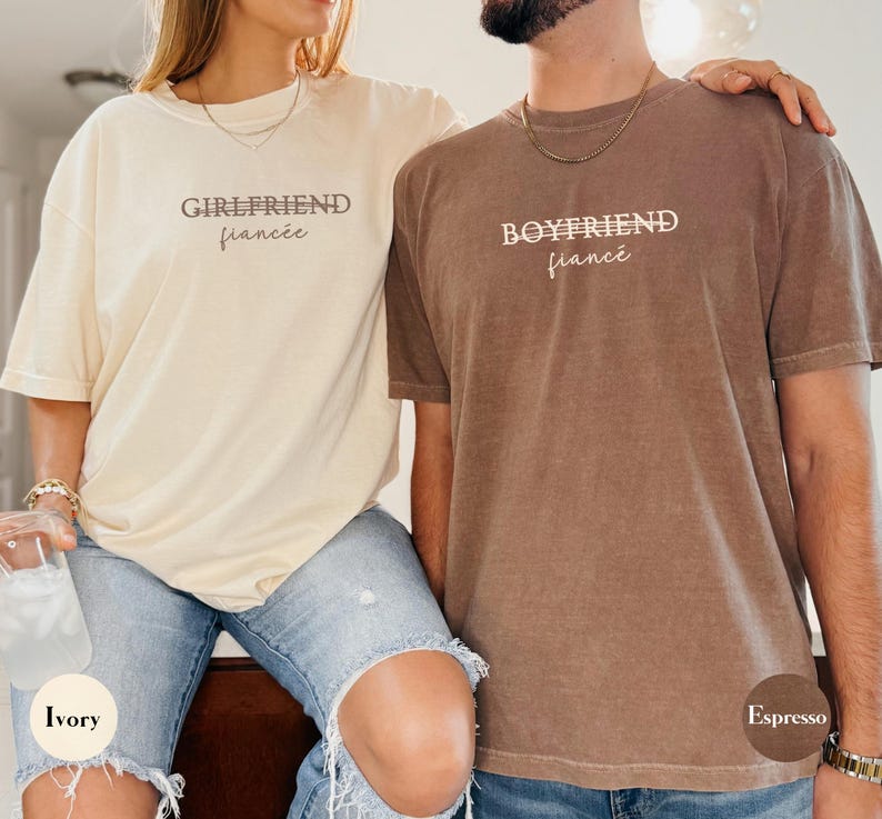 May include: Two matching t-shirts, one ivory and one espresso, with the words "GIRLFRIEND fianc&eacute;e" and "BOYFRIEND fianc&eacute;e" printed on them. The shirts are worn by a couple, suggesting a relationship theme.