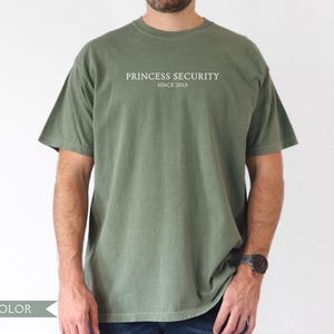 May include: Moss green t-shirt with the text "PRINCESS SECURITY SINCE 2015" in white. The short-sleeved shirt is a solid color and has a relaxed fit. The person is wearing a watch on their left wrist.