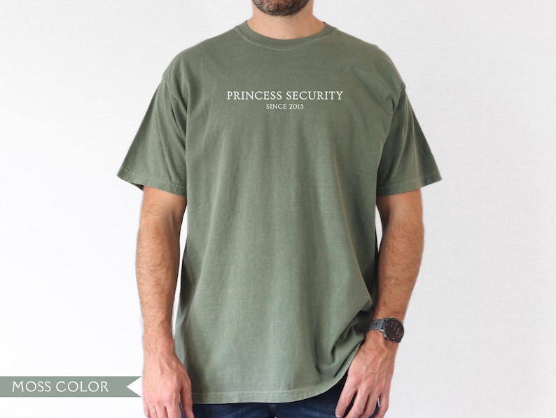 May include: Moss green t-shirt with the text "PRINCESS SECURITY SINCE 2015" in white. The short-sleeved shirt is a solid color and has a relaxed fit. The person is wearing a watch on their left wrist.