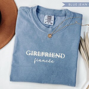 May include: A blue jean colored Comfort Colors t-shirt with the words "GIRLFRIEND" and "fianc&eacute;e" printed in white. A gold necklace with a pendant is draped across the shirt. A brown hat and dried plant are in the background.