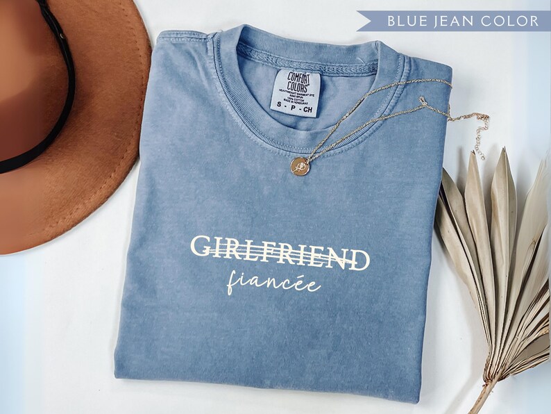 May include: A blue jean colored Comfort Colors t-shirt with the words "GIRLFRIEND" and "fianc&eacute;e" printed in white. A gold necklace with a pendant is draped across the shirt. A brown hat and dried plant are in the background.