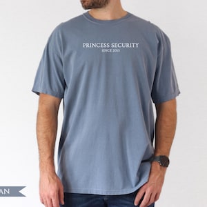 May include: Blue jean colored t-shirt with the text "PRINCESS SECURITY SINCE 2013" in white. The shirt has short sleeves and a crew neck. The model is wearing a black watch.