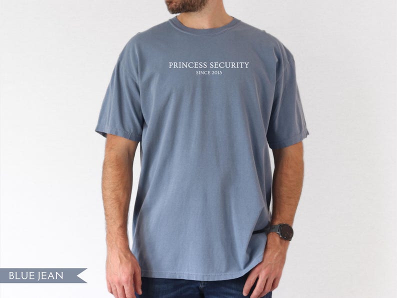 May include: Blue jean colored t-shirt with the text "PRINCESS SECURITY SINCE 2013" in white. The shirt has short sleeves and a crew neck. The model is wearing a black watch.