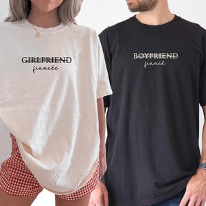 May include: Two t-shirts, one ivory and one black, with the words "GIRLFRIEND fianc&eacute;e" and "BOYFRIEND fianc&eacute;" printed on the front. The ivory shirt is paired with red and white checkered shorts. The black shirt is paired with blue jeans.