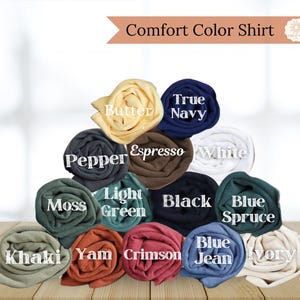 May include: A display of rolled Comfort Color shirts in various colors, including Butter, True Navy, Pepper, Espresso, White, Moss, Light Green, Black, Blue Spruce, Khaki, Yam, Crimson, Blue Jean, and Ivory. The text "Comfort Color Shirt" is at the top.