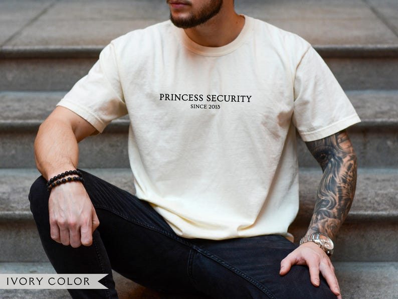 May include: Ivory-colored t-shirt with the text "PRINCESS SECURITY SINCE 2013" in black. The shirt is worn with black jeans. The image also shows a wrist watch and a bracelet.