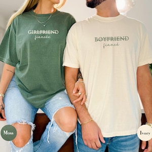 May include: Two matching short-sleeve t-shirts, one moss green and one ivory. The moss green shirt has the text "GIRLFRIEND fianc&eacute;e" in white. The ivory shirt has the text "BOYFRIEND fianc&eacute;e" in a muted green.