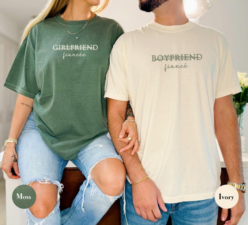 May include: Two matching short-sleeve t-shirts, one moss green and one ivory. The moss green shirt has the text "GIRLFRIEND fianc&eacute;e" in white. The ivory shirt has the text "BOYFRIEND fianc&eacute;e" in a muted green.