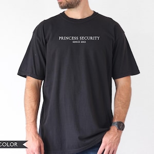 May include: Black t-shirt with the text "PRINCESS SECURITY SINCE 2013" in white. The shirt is a solid black color and has short sleeves. The model is wearing a watch on their left wrist.