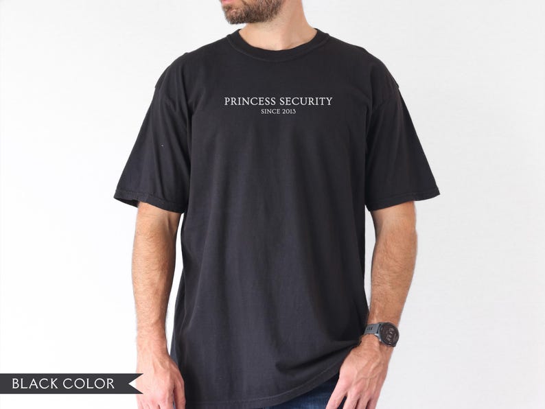 May include: Black t-shirt with the text "PRINCESS SECURITY SINCE 2013" in white. The shirt is a solid black color and has short sleeves. The model is wearing a watch on their left wrist.