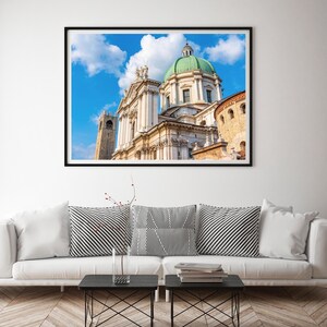 May include: A framed print of a cathedral with a green dome and white facade against a blue sky with clouds. The print is displayed above a white sofa with black and white striped pillows and two small black tables.