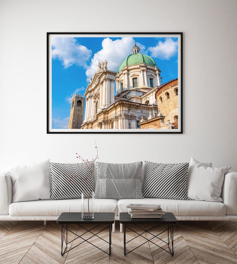 May include: A framed print of a cathedral with a green dome and white facade against a blue sky with clouds. The print is displayed above a white sofa with black and white striped pillows and two small black tables.