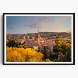 May include: A framed print depicting a European town at sunset. The image shows a tall church spire, terracotta rooftops, and bright yellow trees in the foreground. The sky displays a blend of blue, orange, and pink colours.