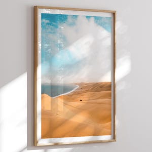 Peut inclure: Une impression encadrée représentant un paysage côtier. L'image montre une plage avec des dunes de sable dorées rencontrant l'océan turquoise sous un ciel bleu nuageux. L'œuvre est encadrée dans un cadre en bois clair.