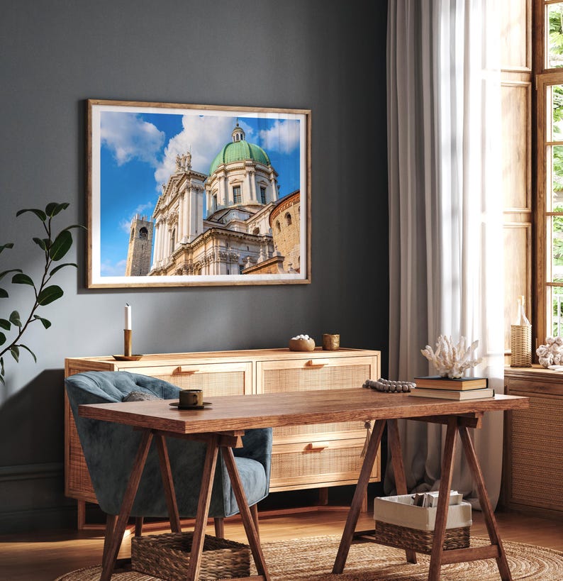 May include: A framed print of a cathedral with a green dome and white facade hangs above a wooden desk and cabinet. The room has a blue chair, a candle, and a window with a white curtain. The image is a photograph.