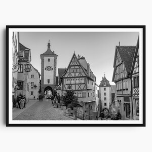 Könnte beinhalten: Schwarz-Weiß-Druck eines europäischen Stadtplatzes mit Kopfsteinpflasterstraßen und historischen Gebäuden. Ein hoher Uhrturm steht in der Mitte, mit mehreren Personen, die die Straße entlanggehen. Die Gebäude haben steil abfallende Dächer.