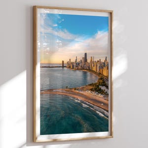 Op de afbeelding: Een ingelijste print met een schilderachtig luchtfoto van de skyline van Chicago langs Lake Michigan. De afbeelding toont de wolkenkrabbers van de stad, een zandstrand en het blauwe water onder een gedeeltelijk bewolkte hemel. Het frame is gemaakt van lichtgekleurd hout.