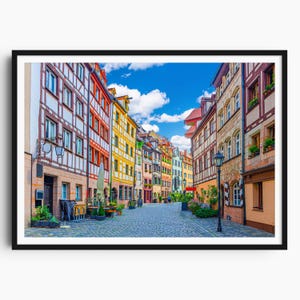 Könnte beinhalten: Ein gerahmter Druck einer europäischen Straßenszene. Bunte Gebäude mit roten, gelben und weißen Fassaden säumen eine Kopfsteinpflasterstraße. Ein strahlend blauer Himmel mit flauschigen weißen Wolken füllt den Hintergrund.
