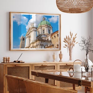 May include: A framed photograph of a cathedral with a green dome and a blue sky. The image is displayed above a wooden cabinet with woven rattan doors. A dining table and chairs are in the foreground, with decorative items on the cabinet.