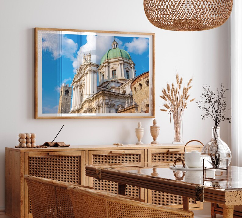 May include: A framed photograph of a cathedral with a green dome and a blue sky. The image is displayed above a wooden cabinet with woven rattan doors. A dining table and chairs are in the foreground, with decorative items on the cabinet.