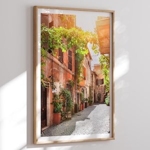 Puede incluir: Una impresión enmarcada de una estrecha calle adoquinada en una ciudad italiana. Los edificios son de color naranja cálido y rojo, con vides y plantas verdes. La luz del sol crea una atmósfera brillante y acogedora.