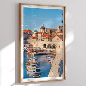 May include: A framed print of a coastal town with red-tiled roofs, a harbor filled with boats, and a clear blue sky. The image captures a sunny day with a view of buildings and a church.