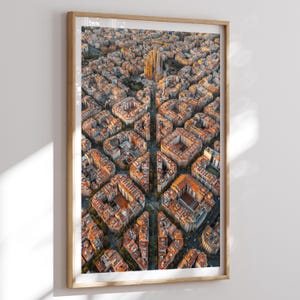 May include: An aerial photograph of a city, framed in light wood. The image shows a dense urban landscape with buildings of varying heights and orange rooftops. A prominent church spire rises in the distance, with a grid-like street layout.
