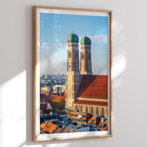 Pode incluir: Impressão emoldurada da Frauenkirche em Munique, Alemanha. A imagem apresenta as torres gêmeas da igreja com cúpulas verdes, um telhado de telhas vermelhas e uma paisagem urbana sob um céu azul. A impressão está em uma moldura de madeira clara.