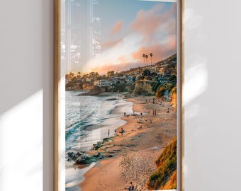 May include: Framed print of a beach scene at sunset. The image features a sandy beach, ocean waves, and a hillside with buildings and palm trees. The sky is filled with pink and orange clouds, creating a warm, inviting atmosphere.