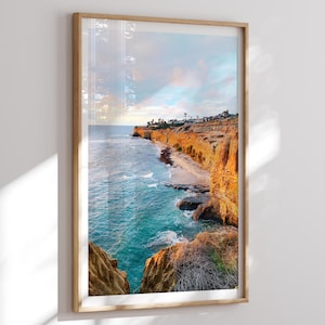 May include: A framed print depicting a coastal landscape. The image shows a cliffside with golden-orange rock formations, turquoise ocean water, and a sandy beach. The sky is a mix of blue and pastel colours.