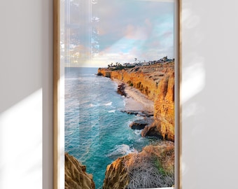 May include: A framed print of a coastal landscape. The image features a vibrant scene of a cliffside with golden-orange rock formations, turquoise ocean water, and a sandy beach. The sky is a mix of blue and pastel colors.