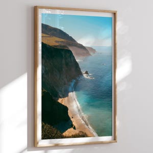 May include: A framed landscape print featuring a coastal scene. The image showcases a beach with turquoise water, cliffs, and mountains under a blue sky. The print is in a wooden frame, adding a natural touch to the artwork.