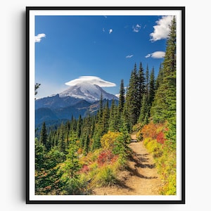Op de afbeelding: Een ingelijste print van een berglandschap. Een pad slingert door een bos van groene en rode bomen, dat leidt naar een besneeuwde berg onder een blauwe lucht met wolken. De print zit in een zwart frame.