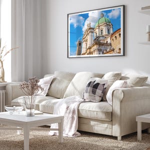 May include: A framed print of a cathedral with a green dome and white facade hangs above a beige sofa. The room features a white coffee table, a patterned pillow, and a light-colored rug, creating a bright and airy living space.