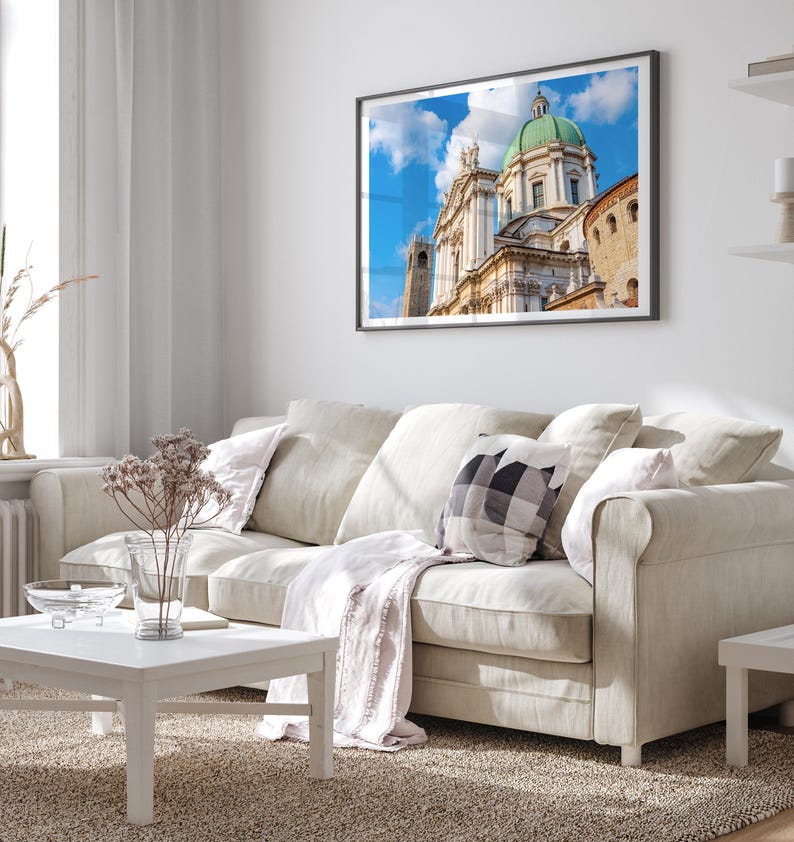 May include: A framed print of a cathedral with a green dome and white facade hangs above a beige sofa. The room features a white coffee table, a patterned pillow, and a light-colored rug, creating a bright and airy living space.