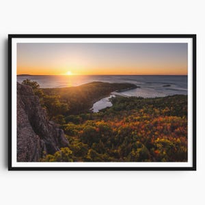 May include: A framed landscape print featuring a vibrant sunset over a coastal scene. The image captures a body of water, a peninsula, and a forest with autumn foliage. The sky transitions from orange to blue, with the sun's rays illuminating the scene.