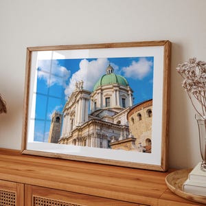May include: A framed print of a grand cathedral with a green dome and white facade against a bright blue sky with fluffy white clouds. The print is in a light brown wooden frame, resting on a wooden cabinet.