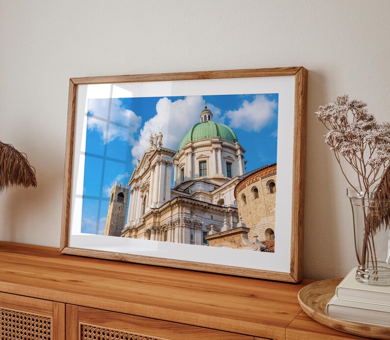 May include: A framed print of a grand cathedral with a green dome and white facade against a bright blue sky with fluffy white clouds. The print is in a light brown wooden frame, resting on a wooden cabinet.