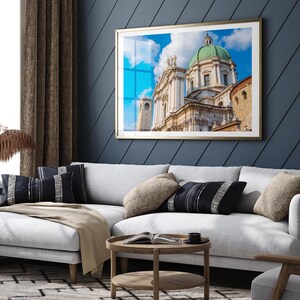 May include: A framed print of a cathedral with a green dome and white facade hangs above a white sectional sofa. The print has a gold frame and is set against a dark blue wall. Decorative pillows and a wooden coffee table complete the living room setting.
