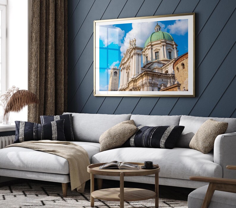 May include: A framed print of a cathedral with a green dome and white facade hangs above a white sectional sofa. The print has a gold frame and is set against a dark blue wall. Decorative pillows and a wooden coffee table complete the living room setting.
